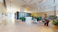 Located in Miami's Wynwood-adjacent neighborhood, this ground-floor lobby presents a sophisticated and welcoming arrival experience. The space is defined by soaring ceilings, large-format stone tile flooring, and floor-to-ceiling glazing that floods the interior with abundant natural light while framing views of the surrounding streetscape. A prominent curved reception desk clad in dark paneling serves as the focal point, flanked by oversized potted tropical plants that reinforce the lush, biophilic atmosphere. Warm light wood paneling lines the walls and elevator corridors, providing a natural contrast to the sleek matte-dark architectural elements. Multiple seating areas are thoughtfully arranged throughout, including houndstooth-upholstered lounge chairs grouped around low coffee tables on richly colored area rugs, inviting casual gathering and relaxation. Curated artwork is displayed prominently throughout, including a vibrant large-scale painting depicting a tropical scene, a colorful abstract canvas near the reception, and sculptural hanging installations visible toward the lounge end of the lobby. A dedicated mail and package room offers organized resident services with an extensive bank of private mailboxes. The overall ambiance is contemporary yet warm, balancing refined design details with a welcoming, resort-inspired sensibility.