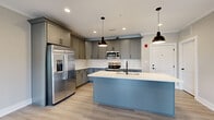 Located at 18442 Leesville Road in Lynchburg, Virginia, this thoughtfully designed single-story home offers three bedrooms, two full bathrooms, and a comfortable, functional layout spread across a generously sized floor plan. The open-concept kitchen and living area serves as the heart of the home, showcasing gray shaker-style cabinetry, crisp white quartz countertops, a subway tile backsplash, and a center island with a built-in sink. Stainless steel appliances, including a French door refrigerator, electric range, and built-in microwave, complete the kitchen. Stylish matte black pendant lights hang above the island, adding a modern touch. Light hardwood-style flooring flows throughout the main living areas, while ceiling fans provide comfort overhead. The spacious primary bedroom features a ceiling fan with natural light from a bright window. Additional bedrooms offer soft carpet, neutral tones, and easy closet access via three dedicated closets throughout the home. Both bathrooms are finished with gray vanities, white quartz countertops, and tub-shower combinations with clean chrome fixtures. A dedicated laundry room houses side-by-side washer and dryer units. French doors in the living area invite natural light and outdoor access, rounding out this move-in-ready home.