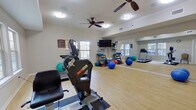 This single-floor fitness center is thoughtfully designed to support a wide range of exercise activities. The space features light hardwood flooring throughout, creating a clean and energizing atmosphere. Multiple windows along the walls allow generous natural light to fill the room, complementing the bright overhead flush-mount lighting. Dual ceiling fans help maintain comfortable airflow during workouts.

The facility is equipped with a variety of cardio machines, including a Matrix treadmill, a ScieFit recumbent cycle, and an elliptical-style stepper. A large wall-to-wall mirror on one side of the room visually expands the space and is ideal for monitoring form during exercise. A wall-mounted flat-screen television provides entertainment during workouts.

The free-weight and functional fitness area includes a well-stocked dumbbell rack with a full range of color-coded hand weights, exercise mats, and an assortment of stability and medicine balls in various sizes. A water dispenser is conveniently positioned within the space for hydration. The room also features a dedicated open-floor zone suitable for stretching, yoga, or group fitness activities. Interior access doors connect to adjacent areas of the facility, making this fitness center a practical and accessible amenity.
