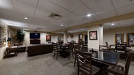 Located at 115 W Monument Ave in Dayton, Ohio, this single-floor community lounge offers a generously sized, open layout designed for socializing and relaxation. The space is divided into several distinct functional zones, all unified by neutral warm-toned walls, drop tile ceilings with recessed lighting, and carpeted flooring throughout.

The lounge area anchors the space with rich brown leather sofas, a loveseat, and an armchair arranged around a classic wood-mantled fireplace topped with a wall-mounted flat-screen television — creating an inviting atmosphere for gathering. Distinctive arched leaded-glass windows add an elegant architectural accent to this seating nook, complemented by decorative artwork and a tall indoor plant.

Adjacent to the lounge, multiple dining and game tables with sturdy dark wood chairs provide ample seating for group activities. A high-top bar counter running along one wall is lined with stylish metal barstools with upholstered seats, offering additional casual seating.

A well-equipped kitchenette supports the space with light wood cabinetry, a double sink, microwave, dishwasher, and a full-size refrigerator — ideal for events and gatherings. The overall ambiance is warm, comfortable, and functional, making this a versatile space well-suited for community events, dining, and leisure activities.