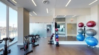 This contemporary fitness center is designed to support a full range of workout activities within a clean, modern environment. The space is thoughtfully organized into distinct functional zones, separated by frosted glass partitions that provide visual privacy while maintaining an open, airy feel throughout.

The main workout area features hardwood-style flooring and is outfitted with stationary cycling bikes, ideal for cardio training. A dedicated strength and functional training zone houses a well-stocked medicine ball rack with a variety of weighted balls, catering to diverse fitness routines. A floor-to-ceiling mirror lines one wall, enhancing the sense of space and allowing users to monitor their form during exercise.

Recessed linear LED lighting runs along the white ceilings, providing bright, even illumination throughout the facility. A secondary area visible through the glass partition appears to accommodate additional equipment and training space, expanding the overall capacity of the center.

The overall ambiance is professional and motivating, combining a neutral color palette of whites and grays with warm flooring tones. The space is well-ventilated and thoughtfully laid out, making it a functional and inviting environment for a wide range of fitness activities.