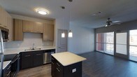 This single-floor space showcases a contemporary open-concept layout that seamlessly connects the kitchen and living areas. The kitchen is equipped with dark-toned cabinetry paired with light countertops, creating a striking contrast. A central island with matching dark cabinetry and a light quartz-style surface serves as a functional focal point, illuminated by two stylish pendant lights with brushed gold accents. Stainless steel appliances, including a dishwasher, complement the modern aesthetic, while light wood upper cabinets add a warm, balanced touch. The sink area is neatly positioned along the perimeter cabinetry. Hardwood-style flooring flows continuously from the kitchen into the open living area, enhancing the sense of cohesion and space. The living area benefits from natural light streaming through a large window fitted with horizontal blinds, and a ceiling fan ensures year-round comfort. Neutral gray walls throughout contribute to a clean, understated palette. A paneled interior door with a gray finish adds a subtle architectural detail, while flush and pendant lighting provide layered illumination across the entire space.