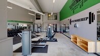 Located at 1505 E Burnsville Pkwy in Burnsville, Minnesota, this single-floor fitness center offers a comprehensive workout environment designed to motivate and accommodate a wide range of exercise needs. The space features soaring ceilings with exposed ductwork, lending an open, industrial feel that keeps the atmosphere energetic and spacious.

The main workout floor is generously sized and thoughtfully divided into distinct training zones. A robust selection of strength machines — including cable systems, leg press equipment, and various selectorized units — is arranged throughout the space, complemented by stability balls and a rowing machine for functional fitness. A dedicated cardio zone lines the perimeter, where upright bikes, recumbent bikes, and treadmills are positioned directly in front of expansive floor-to-ceiling windows that bathe the area in natural light and offer views of the outdoor pool and surrounding greenery.

Bold motivational graphics in green and black adorn the walls, including a large fitness-themed word mural and the "ONEMOREREP" branding, reinforcing the facility's energetic identity. Mirrored wall sections enhance the sense of depth and allow users to monitor their form during workouts. The flooring transitions between polished concrete and rubber mat sections, providing both durability and safety. Overall, the space delivers a bright, modern, and fully equipped fitness experience.