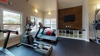 Located at 997 Johnnie Dodds Blvd in Mount Pleasant, South Carolina, this well-equipped fitness center offers a comprehensive workout environment within a single-floor layout. The space is defined by its dramatic vaulted ceilings, which lend an open, airy atmosphere and amplify the sense of spaciousness throughout. Large windows line the walls, allowing generous natural light to fill the room while offering views of the surrounding greenery.

The cardio zone features multiple Star Trac treadmills and elliptical machines, arranged to face the windows and a wall-mounted flat-screen television set against a striking wood-panel accent wall. Built-in cubbies beneath the TV provide organized storage for exercise accessories, including stability balls and resistance equipment.

The strength training area is equally well-appointed, housing a Matrix multi-functional cable machine, a power tower for bodyweight exercises, and a full rack of dumbbells spanning a wide weight range. Floor-to-ceiling mirrors line the walls, enhancing the sense of depth and allowing users to monitor their form throughout their workouts.

The durable rubber flooring provides safety and cushioning across the entire space. Recessed lighting and wall sconces complement the natural light, ensuring the facility is well-lit at any hour. French doors offer direct outdoor access, further connecting the interior to the pleasant South Carolina surroundings.