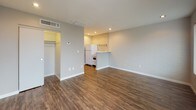 This single-floor unit offers a practical and well-organized layout designed to maximize every inch of space. The main living area is open and airy, finished with rich wood-look vinyl flooring that runs throughout and painted brick-accent walls that lend a subtle industrial character to the interior. Recessed lighting illuminates the space evenly, while a large window fitted with vertical blinds allows natural light to flow in alongside a glass-paneled entry door.

The kitchen is efficiently arranged and directly accessible from the living area via a peninsula counter. It is equipped with white appliances including a full-size refrigerator, a four-burner electric coil range with oven, and a wall-mounted utility shelf above. White subway tile backsplash and light-toned countertops complete the clean, functional aesthetic.

Off the main living space, a generously sized walk-in closet provides ample storage with dual hanging rods mounted along painted brick walls and matching wood-look flooring. The bathroom is neatly appointed with light tile flooring, white brick-style wall tile, and a full toilet and bath setup. A utility panel is conveniently located in the hallway just outside the bathroom, keeping essential systems accessible.