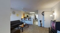 This two-story residence features an open-concept main living area that seamlessly connects the living room and kitchen, creating a spacious, cohesive environment. The living room is carpeted and furnished with a dark sofa and a wooden coffee table, with a ceiling fan overhead providing both airflow and casual lighting. Wall décor, including a sunburst clock and a framed accent piece, adds character to the neutral-toned walls.

The kitchen is positioned adjacent to the living area and is equipped with white cabinetry, a black microwave, a full-size refrigerator, and under-cabinet fluorescent lighting. A raised breakfast bar with bar-height seating for three separates the kitchen from the living space, making it ideal for casual dining or entertaining. The kitchen flooring transitions from the carpeted living area to a hard surface, clearly delineating the two zones.

A hallway visible from the living area leads to additional rooms, including what appears to be a home office or study space. The overall layout is practical and well-organized, with the two-floor design offering additional living or sleeping space beyond the open main level.