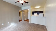 This single-floor space features an open-concept design that connects the living area to the kitchen, creating a functional and cohesive layout. The living area is finished with neutral-toned carpet, providing a comfortable underfoot feel, while a ceiling fan with a built-in light fixture serves as both a practical and decorative element overhead. Warm, neutral-toned walls contribute to an inviting atmosphere throughout the space.

The kitchen is positioned adjacent to the living area, separated by a raised breakfast bar counter that doubles as a casual dining or workspace surface. Dark espresso-toned cabinetry lines the upper kitchen walls, offering a striking contrast against the lighter walls and countertops. A white refrigerator and a pendant light fixture are also visible, adding functional character to the kitchen zone.

A hallway visible from the living area leads further into the space, where hardwood-style flooring transitions from the carpeted areas. Through an open doorway at the end of the hall, a laundry area with stacked or side-by-side appliances can be seen, indicating convenient in-unit laundry access. The overall layout feels practical and well-organized.