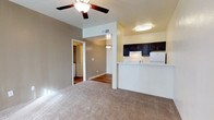This single-floor space features an open-concept design that connects the living area to the kitchen, creating a functional and cohesive layout. The living area is finished with neutral-toned carpet, providing a comfortable underfoot feel, while a ceiling fan with a built-in light fixture serves as both a practical and decorative element overhead. Warm, neutral-toned walls contribute to an inviting atmosphere throughout the space.

The kitchen is positioned adjacent to the living area, separated by a raised breakfast bar counter that doubles as a casual dining or workspace surface. Dark espresso-toned cabinetry lines the upper kitchen walls, offering a striking contrast against the lighter walls and countertops. A white refrigerator and a pendant light fixture are also visible, adding functional character to the kitchen zone.

A hallway visible from the living area leads further into the space, where hardwood-style flooring transitions from the carpeted areas. Through an open doorway at the end of the hall, a laundry area with stacked or side-by-side appliances can be seen, indicating convenient in-unit laundry access. The overall layout feels practical and well-organized.