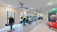 This fitness center offers a clean, modern environment designed to support a comprehensive workout experience. The space is finished with light hardwood-style flooring and crisp white walls that contribute to an open, energizing atmosphere. Large windows line the walls, flooding the interior with natural light and providing views of the surrounding greenery, while recessed lighting ensures consistent illumination throughout. Ceiling fans are strategically positioned overhead, promoting airflow and comfort during workouts. A full-length mirror along one wall visually expands the space and assists users in monitoring their form. The cardio zone is well-appointed with a row of treadmills and elliptical machines from TRUE Fitness, alongside a recumbent bicycle, offering a variety of aerobic options. A dedicated free-weight and functional training area includes a tiered dumbbell rack and a selection of stability balls in multiple sizes displayed on a standing rack, along with exercise mats for floor-based training. The layout is thoughtfully organized to allow free movement between equipment zones. With its bright finishes, quality equipment, and practical design, this fitness center provides a welcoming and functional environment for daily exercise.