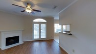 This two-story home opens into a welcoming main living area distinguished by warm hardwood-style flooring and classic crown molding that frames the ceiling throughout. The focal point of the entry is a set of elegant French doors topped with an arched transom window, allowing generous natural light to flood the interior and creating an inviting, airy atmosphere. A ceiling fan with a light fixture is centrally positioned overhead, providing both comfort and functionality. Large windows along the adjacent wall further enhance the brightness of the space. A half-wall partition with a dark countertop surface separates the main living area from a neighboring room, hinting at an open-concept layout that promotes connectivity between spaces. The adjoining area features tile flooring, offering a practical transition zone, likely leading toward a kitchen or dining area. Neutral wall tones throughout complement the light-colored trim and baseboards, lending the home a clean, versatile aesthetic. The combination of architectural detailing, abundant natural light, and thoughtful layout makes this a functional and visually appealing two-story home.