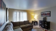 This single-floor residence offers a welcoming living area that serves as the focal point of the space. The living room features neutral-toned walls that create a warm, inviting atmosphere, complemented by smooth, light-colored ceilings that enhance the sense of openness. A large window dressed with vertical blinds allows natural light to filter into the room, brightening the carpeted floor and highlighting the comfortable sectional sofa positioned as the room's centerpiece. A small side table with a lamp provides additional warm lighting, contributing to the cozy ambiance. Dark-finished furniture pieces, including a multi-drawer dresser, add contrast against the lighter walls and flooring. Wall art adds a decorative touch to the space, while a woven area rug anchors the seating arrangement. The overall design is practical and functional, with a straightforward layout that maximizes the available floor space. The neutral color palette throughout creates a cohesive, understated aesthetic that offers a comfortable and livable environment suitable for everyday use.