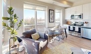 This well-designed single-floor residence offers a clean, contemporary aesthetic with light-toned walls, light wood flooring, and recessed lighting throughout. A welcoming hallway with crisp white walls and recessed ceiling lights leads into the main living areas.

The open living room is a standout space, bathed in abundant natural light from large windows that frame elevated outdoor views of the surrounding landscape. The room comfortably accommodates both a seating area and a dining nook, making it ideal for everyday living and entertaining.

The kitchen is thoughtfully designed with white shaker-style cabinetry extending to the ceiling, granite countertops, a subway tile backsplash, a stainless steel undermount sink, and stainless steel appliances including a refrigerator and dishwasher.

Two bedrooms provide comfortable retreats, each featuring light neutral walls and carpet or wood-look flooring. One bedroom benefits from a large window with scenic outdoor views, filling the space with natural light.

The full bathroom is finished with white cabinetry, a single vanity with a white countertop, a combination tub and shower, and warm wood-look flooring, maintaining the clean and cohesive design found throughout the residence. Overall, this space presents a bright, functional, and modern living environment.