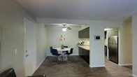 This single-floor space showcases an open-concept layout that seamlessly connects the dining area and kitchen. The dining space is anchored by a round white table surrounded by sleek, modern chairs with chrome legs, positioned beneath a ceiling fan that provides both functionality and a casual aesthetic. Neutral white walls provide a clean backdrop, complemented by a colorful piece of wall art that adds a pop of personality to the space.

The adjacent kitchen features dark lower cabinetry and upper cabinets in a contrasting tone, paired with a light countertop that runs along the perimeter. A smart thermostat is mounted on the wall, indicating modern technological integration throughout the space. The kitchen is well-equipped, with appliances visible in the background, and the layout allows for an efficient workflow.

Rich, dark hardwood-style flooring flows continuously throughout both areas, creating a cohesive and grounded feel. The overall ambiance is contemporary and understated, with a neutral color palette and modern furnishings that make the space feel both functional and inviting. The open layout enhances the sense of spaciousness while maintaining a clear distinction between living zones.