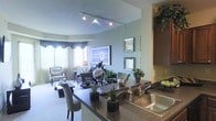 This single-floor residence showcases a thoughtfully designed open-concept layout that connects the kitchen, dining, and living areas into one cohesive, flowing space. The kitchen features dark granite-style countertops, a stainless steel sink, and warm wood cabinetry that extends to the ceiling, providing generous storage. A peninsula counter with seating bridges the kitchen and dining area, making it ideal for casual dining and entertaining. The living area is anchored by plush carpeting and bathed in soft, ambient light from a sleek track lighting system mounted to the ceiling. Large windows dressed with elegant draping curtains allow natural light to filter into the space, creating a bright and airy atmosphere. Decorative mirrors and framed wall art add visual depth and personality to the soft sage-green walls. Touches of greenery throughout — from potted plants on the counter to tall arrangements atop the cabinetry — lend a fresh, organic quality to the interior. The overall ambiance is warm, modern, and welcoming, with a careful balance of functional design and refined aesthetic details that make this space both comfortable and visually appealing.