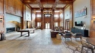 This expansive single-floor lodge-style facility makes an immediate impression with its grand entrance corridor, featuring natural stone tile flooring, rich knotty wood wall paneling, and soaring double-height ceilings that flood the space with natural light through tall clerestory windows. The main gathering hall is a showstopper — anchored by an oversized chandelier and framed by towering wood-clad columns, it seamlessly combines a billiards area, multiple seating vignettes with leather and upholstered furnishings, and a stone-surround fireplace, all set against warm neutral walls and rustic stone tile floors. Multiple lounge areas branch off the central space, each thoughtfully appointed with curated artwork and accent lighting, creating intimate settings within the larger open layout. Elegant wrought iron railings delineate seating nooks near large windows overlooking mature trees outside. French doors with multi-pane glass panels connect the various zones, allowing natural light to flow throughout while maintaining a sense of defined purpose for each area. A dedicated business or leasing office area features built-in wood millwork and workstation furnishings. The cohesive use of knotty wood, stone, and warm lighting throughout lends the entire space a refined yet welcoming mountain-lodge aesthetic.
