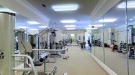 This fitness center offers a spacious and well-organized workout environment designed to accommodate a wide range of exercise needs. The layout is thoughtfully arranged, with a variety of strength-training machines and cable systems positioned throughout the room, providing ample space for users to move freely between equipment. The flooring consists of a neutral, low-pile carpet that spans the entire area, offering both comfort and stability underfoot. Floor-to-ceiling mirrors line one full wall, creating a sense of depth and allowing users to monitor their form during workouts. The space is bathed in bright, even light from large flush-mounted overhead fixtures, ensuring excellent visibility throughout. Crown molding along the upper walls and neutral, warm-toned paint lend the space a polished, upscale aesthetic that sets it apart from a typical gym environment. A dumbbell and free weight rack is visible toward the far end of the room, alongside additional cable and resistance machines. Natural light is supplemented by the artificial lighting, contributing to an open and energizing atmosphere. The overall ambiance is clean, professional, and inviting, making it a functional and comfortable space for daily fitness routines.