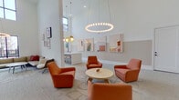 This single-floor space is designed as a sophisticated communal lounge, distinguished by dramatically high ceilings that lend an airy, open quality throughout. The layout is divided into several functional zones that flow seamlessly into one another, unified by a cohesive neutral palette accented with warm terracotta and earthy tones.

The main lounge area centers around a grouping of bold orange upholstered armchairs arranged around a round light wood coffee table, anchored by a patterned area rug. A striking circular halo pendant light hangs overhead, complementing additional sculptural gold disc pendants nearby. Curated framed artwork in soft blush and desert-inspired tones lines the walls, adding visual depth.

A dedicated workspace features a long white communal table lined with tan bar stools, illuminated by gold pendant lights and bathed in natural light from expansive floor-to-ceiling black-framed windows that provide outdoor views.

An arched accent wall clad in a multicolored mosaic tile serves as a standout architectural feature, framing a built-in white credenza beneath. Adjacent seating includes a neutral sofa and lounge chair, completing the space's relaxed yet refined ambiance.