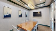 Located at 1055 Pine St in Nashville, TN, this compact yet well-appointed conference room offers a refined setting for professional meetings and collaborative sessions. The room centers around a sleek hardwood conference table with a glossy finish, surrounded by six cushioned chairs in a soft, neutral tone — comfortably accommodating a small group for focused discussions. A large wall-mounted flat-screen display serves as a key feature, providing a dedicated surface for presentations and media sharing. The ceiling draws immediate attention with its distinctive layered tray design, accented by deep teal and cream striped detailing, and anchored by a statement cylindrical pendant light that adds warmth and a contemporary flair. The walls are finished with a textured grasscloth-style wallcovering in soft gray, complemented by a curated set of framed abstract artwork in blue and neutral tones. Teal baseboards and trim elements tie the color palette together cohesively throughout the space. Glass panel walls and a glass-paned door on one side provide visual connectivity to an adjacent common area, lending an open and professional atmosphere. Carpeted flooring in a warm, speckled tone completes the interior, balancing the overall design with a grounded, functional aesthetic.