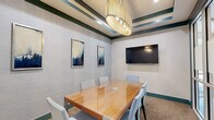 Located at 1055 Pine St in Nashville, TN, this compact yet well-appointed conference room offers a refined setting for professional meetings and collaborative sessions. The room centers around a sleek hardwood conference table with a glossy finish, surrounded by six cushioned chairs in a soft, neutral tone — comfortably accommodating a small group for focused discussions. A large wall-mounted flat-screen display serves as a key feature, providing a dedicated surface for presentations and media sharing. The ceiling draws immediate attention with its distinctive layered tray design, accented by deep teal and cream striped detailing, and anchored by a statement cylindrical pendant light that adds warmth and a contemporary flair. The walls are finished with a textured grasscloth-style wallcovering in soft gray, complemented by a curated set of framed abstract artwork in blue and neutral tones. Teal baseboards and trim elements tie the color palette together cohesively throughout the space. Glass panel walls and a glass-paned door on one side provide visual connectivity to an adjacent common area, lending an open and professional atmosphere. Carpeted flooring in a warm, speckled tone completes the interior, balancing the overall design with a grounded, functional aesthetic.