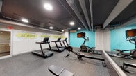 This fitness center offers a focused and functional workout environment designed to accommodate a variety of exercise routines. The space is laid out on a single level with durable rubber-speckled flooring throughout, providing both safety and comfort underfoot during workouts.

The cardio zone is anchored by multiple treadmills lined along one wall, complemented by an elliptical trainer and a HIIT rowing machine, offering users a well-rounded selection of cardiovascular equipment. A wall-mounted flat-screen television faces the cardio area, positioned against a bold teal accent wall that adds a vibrant visual focal point to the room.

A dedicated strength training area features a multi-tiered dumbbell rack stocked with a range of free weights, alongside a flat workout bench and a core exercise mat station. A large wall mirror spans one side of the space, visually expanding the room and allowing users to monitor their form during exercise.

The exposed ceiling with visible ductwork and overhead recessed lighting gives the space an industrial aesthetic, while an in-wall air conditioning unit ensures climate comfort. Energetic wall murals featuring fitness-themed typography—highlighting words such as "Nutrition," "Fitness," "Training," and "Attitude"—contribute to a motivating atmosphere. An exit and a private door provide convenient access points throughout the facility.