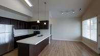 This single-floor space showcases a thoughtfully designed open-concept layout that seamlessly connects the kitchen and living areas. The kitchen is a standout feature, outfitted with rich dark espresso cabinetry that contrasts elegantly against crisp white countertops. A peninsula with an integrated sink serves as both a functional workspace and a natural divider between the kitchen and the adjoining living area. Black appliances, including a range and built-in microwave, add a cohesive modern aesthetic. Pendant lighting suspended from the vaulted ceiling casts warm, focused light over the peninsula, while a skylight overhead floods the kitchen with natural daylight. The living area benefits from soaring vaulted ceilings, amplifying the sense of openness and volume throughout the space. Track lighting installed along the upper wall provides flexible, adjustable illumination for the living zone. Smooth, light-toned hardwood-style flooring runs continuously throughout, unifying the open floor plan and adding warmth underfoot. Crisp white walls and clean trim work keep the overall palette bright and neutral, allowing the dark cabinetry and fixtures to serve as bold focal points. A hallway visible in the background suggests additional rooms beyond the main living area.