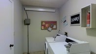 This single-floor laundry facility is designed with functionality and community use in mind. The room features light gray walls and a textured ceiling that contribute to a clean, utilitarian aesthetic. A row of white top-load washers and dryers lines the space, providing ample capacity for shared use. The machines are well-organized and easily accessible, making the laundry process straightforward for residents or guests. A cork bulletin board mounted on the wall serves as a community communication hub, displaying notices and decorative touches that add a welcoming feel. Signage, including a "Laundry" placard and a "Fluff and Fold" wall display, clearly identifies the room's purpose and available services. An electrical panel box is mounted on one wall, reflecting the room's practical infrastructure. The tile flooring is easy to maintain and well-suited for the demands of a high-use laundry space. Overhead fluorescent lighting ensures the room is well-lit throughout. Overall, this laundry facility balances practicality with a modest sense of community character, offering a clean and organized environment for everyday laundry needs.