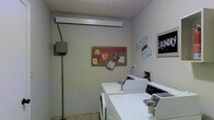 This single-floor laundry facility is designed with functionality and community use in mind. The room features light gray walls and a textured ceiling that contribute to a clean, utilitarian aesthetic. A row of white top-load washers and dryers lines the space, providing ample capacity for shared use. The machines are well-organized and easily accessible, making the laundry process straightforward for residents or guests. A cork bulletin board mounted on the wall serves as a community communication hub, displaying notices and decorative touches that add a welcoming feel. Signage, including a "Laundry" placard and a "Fluff and Fold" wall display, clearly identifies the room's purpose and available services. An electrical panel box is mounted on one wall, reflecting the room's practical infrastructure. The tile flooring is easy to maintain and well-suited for the demands of a high-use laundry space. Overhead fluorescent lighting ensures the room is well-lit throughout. Overall, this laundry facility balances practicality with a modest sense of community character, offering a clean and organized environment for everyday laundry needs.
