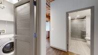 Located in Chicago's dynamic Streeterville neighborhood, this single-floor loft at 445 E Illinois Street masterfully blends industrial character with modern finishes. The open-concept living and kitchen area is anchored by soaring wood-plank ceilings with exposed beams and ductwork, structural timber columns, and gleaming hardwood floors that reflect the warm overhead track lighting. The kitchen is well-appointed with rich walnut cabinetry, granite countertops, a large center island, subway tile backsplash, and a full suite of stainless steel appliances including a gas range and refrigerator. The bedroom spaces showcase original exposed brick walls punctuated by large double-hung windows that flood the rooms with natural light and urban views, while plush carpeting adds warmth underfoot. Ceiling fans provide comfort throughout. The bathroom features a sleek, modern design with a wide white quartz vanity, undermount sink, dark wood cabinetry, and a tiled walk-in shower. A dedicated in-unit laundry area with a front-load washer and built-in shelving adds everyday convenience. Louvered closet doors and thoughtful storage solutions are found throughout. The space strikes a compelling balance between historic loft aesthetics and contemporary living.