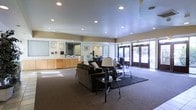 This single-floor commercial space is designed to create a professional and welcoming first impression. The lobby area serves as the heart of the space, featuring a well-appointed reception desk finished in warm wood cabinetry, complete with a large mirror backdrop and organized counter space — ideal for greeting visitors and managing day-to-day front-desk operations. Framed artwork and certificates on the walls add a polished, professional touch.

The lounge area is thoughtfully furnished with plush, dark upholstered sofas arranged around a sleek glass-top coffee table, all anchored by a rich, dark area rug. A pair of classic table lamps provide warm ambient lighting to complement the recessed ceiling lights overhead, which illuminate the space evenly throughout.

Large glass entry doors at the front of the space flood the interior with natural light and offer views of lush greenery outside, creating a bright and open atmosphere. The neutral-toned carpet flooring and light-colored ceiling contribute to a clean, cohesive aesthetic. Overall, this space strikes a balance between functionality and comfort, making it well-suited for a medical office, corporate headquarters, or professional services environment.