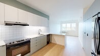Located at 3260 SE Milwaukie Ave in Portland, Oregon, this single-floor 1-bedroom unit offers a thoughtfully designed layout with a clean, modern aesthetic. Light oak hardwood flooring flows throughout the space, complementing the bright white walls and crisp finishes.

The kitchen is a standout feature, showcasing flat-front sage-gray cabinetry extending floor to ceiling, white square-tile backsplash, and white quartz countertops. It is equipped with a stainless steel range, built-in dishwasher, and a French door refrigerator. A peninsula counter seamlessly connects the kitchen to the open living area, enhancing the sense of flow and functionality. Tall upper cabinets and a full-height pantry section provide ample storage.

The living area benefits from a large window that invites natural light and frames views of the surrounding Portland neighborhood. The bedroom is a comfortable size, featuring light-filled windows with treetop views, light hardwood flooring, and a sleek black-hardware sliding barn door that adds a modern industrial touch. A panel for utilities is neatly integrated into the bedroom wall.

The overall design palette — soft greens, whites, and warm wood tones — gives the space a fresh, contemporary character well-suited to urban living in Portland's SE corridor.