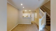 This two-story residence offers a thoughtfully designed layout centered around an open-concept main level. The living area is finished with wall-to-wall carpeting in a warm beige tone, creating a comfortable and cohesive feel throughout the space. Crown molding lines the ceiling, adding a subtle architectural detail that elevates the overall finish of the room.

The kitchen is positioned at the rear of the main level, accessible via a raised peninsula counter with a stone-look surface that provides both workspace and a casual dining option. White cabinetry and white appliances contribute to a clean, classic aesthetic, while recessed lighting above the kitchen and living areas ensures consistent illumination throughout. A door adjacent to the kitchen leads to an additional room or storage area, adding functional versatility to the floor plan.

A white-railed staircase is prominently positioned on the right side of the main level, providing access to the upper floor. The neutral warm-beige wall color flows seamlessly across both levels, reinforcing a unified and welcoming atmosphere throughout the residence.