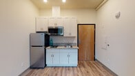 Located at 5603 SE Milwaukie Ave in Portland, Oregon, this single-floor unit offers a functional and efficiently designed layout suited for straightforward, everyday living. The space is finished with light wood-tone luxury vinyl plank flooring that runs consistently throughout, contributing to a clean and cohesive aesthetic.

The kitchenette is thoughtfully outfitted with white shaker-style cabinetry extending to the ceiling, providing ample storage. A granite-patterned countertop houses a stainless steel sink, and stainless steel appliances — including a full-size refrigerator and an over-the-range microwave — complete the cooking area. A warm wood-toned entry door adds a natural contrast to the otherwise neutral palette.

The living and sleeping area is a straightforward, open room with neutral walls and a window fitted with horizontal blinds that allow natural light to filter in while maintaining privacy. The space is versatile enough to accommodate both sleeping and seating arrangements.

The bathroom is ADA-accessible, featuring grab bars, a wall-mounted sink with mirror, and wood-tone flooring that complements the rest of the unit. The overall ambiance is clean, modern, and practical.