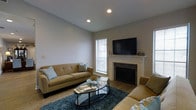 The Mansion is a thoughtfully designed single-floor space that combines comfort and modern style throughout its layout. The living area serves as the heart of the home, showcasing a clean, neutral color palette with light gray walls and white trim that create an open and airy atmosphere. A classic white-mantled fireplace with a dark tile surround anchors the room, topped by a mounted flat-screen television, making it both a functional and stylish focal point. Recessed ceiling lighting provides warm, even illumination throughout the space, complementing the natural light that filters in through a window dressed with horizontal blinds. The living area is furnished with plush tan sofas accented by decorative pillows, arranged around a sleek oval glass coffee table atop a rich teal area rug, adding a pop of color to the otherwise neutral tones. Wood-look flooring runs beneath the furnishings, adding warmth and texture to the space. The overall ambiance is inviting and contemporary, offering a comfortable environment suited for relaxing or entertaining. The cohesive design choices throughout reflect a modern sensibility with timeless touches.