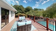This single-story home features a generously sized outdoor deck that serves as a standout extension of the living space. Constructed with rich, warm-toned hardwood planks, the deck provides a polished and durable surface well-suited for both lounging and entertaining. The railing system combines stained wood posts with sleek black metal balusters, offering a refined aesthetic while framing expansive views of the surrounding landscape.

The deck wraps along the exterior of the home, which is clad in crisp white horizontal siding beneath a classic gray asphalt shingle roof. A traditional wall-mounted lantern adds a charming accent near the entrance point. Double-hung windows and a set of dark-framed French doors connect the interior to the deck seamlessly, allowing natural light to flow both ways and creating an easy indoor-outdoor transition.

The outdoor space overlooks a pool and a lush backdrop of mature trees, enhancing the sense of privacy and natural serenity. The deck is furnished with wicker lounge seating accented by aqua cushions and a patterned area rug, reflecting a relaxed, resort-inspired ambiance perfectly suited to the open-air setting.