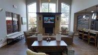 This sophisticated single-floor space is designed to impress with its soaring ceilings and sweeping architectural scale. The focal point of the room is a commanding stone fireplace, crafted from stacked natural stone and topped with a dark wood mantel, above which a large flat-screen television is mounted for added functionality. Flanking the fireplace on either side are tall, arched windows with distinctive grid-paned detailing that stretch toward the ceiling, bathing the interior in abundant natural light and offering pleasant views of the surrounding greenery and outdoor spaces. The seating arrangement is thoughtfully curated, featuring plush upholstered armchairs and sofas in neutral tones that create a warm, welcoming atmosphere. A rustic wooden coffee table serves as the centerpiece of the seating area, anchoring the space with a touch of organic character. Soft table lamps placed symmetrically on either side of the fireplace add a warm ambient glow. The overall ambiance strikes a balance between refined elegance and comfortable functionality, making this a welcoming gathering space well-suited for relaxation and social interaction.