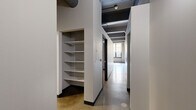 This one-bedroom, one-bathroom loft showcases a distinctive industrial aesthetic defined by exposed ductwork, structural columns, and polished concrete floors throughout the main living areas. The open-concept kitchen serves as a focal point, outfitted with dark cabinetry, granite countertops, stainless steel appliances, and a large island with pendant lighting suspended from the dramatic ceiling above — a design detail that adds both function and visual interest.

The living area benefits from oversized, multi-pane windows that invite generous natural light and offer views of surrounding greenery. The bedroom features plush carpeting, a dedicated closet with hanging rod, and its own access to built-in shelving storage. An additional open-shelf closet space near the entryway corridor provides practical organization solutions.

The bathroom is finished with large-format white wall tiles, a sleek glass-enclosed walk-in shower with a mosaic tile accent band, a dark-cabinetry vanity with a light stone countertop, and polished tile flooring. A utility room and additional closet round out the layout, offering ample storage. The overall ambiance blends urban industrial character with modern finishes, creating a well-appointed and thoughtfully designed living space.