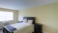 This two-storey residence offers a straightforward and functional layout spread across two levels. The bedroom pictured showcases a clean, minimalist design characterized by soft, neutral wall tones in a warm off-white palette that create a calm and restful atmosphere. Dark-stained wood furnishings, including a classic sleigh-style bed frame and matching nightstands with silver hardware, provide a grounding contrast to the light walls and pale ceiling. The room is comfortably sized, accommodating the bed and flanking nightstands with ease. A window along one wall allows natural light to filter into the space, contributing to the bright and airy feel. Dark hardwood or laminate flooring anchors the room with a rich, warm tone, complementing the dark furniture pieces cohesively. The overall ambiance of the bedroom is tidy and practical, with a traditional design sensibility. White baseboards add a crisp finishing detail throughout. The two-floor layout of the residence suggests additional living, sleeping, or functional spaces distributed between levels, offering a comfortable and organized living environment.