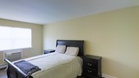 This two-storey residence offers a straightforward and functional layout spread across two levels. The bedroom pictured showcases a clean, minimalist design characterized by soft, neutral wall tones in a warm off-white palette that create a calm and restful atmosphere. Dark-stained wood furnishings, including a classic sleigh-style bed frame and matching nightstands with silver hardware, provide a grounding contrast to the light walls and pale ceiling. The room is comfortably sized, accommodating the bed and flanking nightstands with ease. A window along one wall allows natural light to filter into the space, contributing to the bright and airy feel. Dark hardwood or laminate flooring anchors the room with a rich, warm tone, complementing the dark furniture pieces cohesively. The overall ambiance of the bedroom is tidy and practical, with a traditional design sensibility. White baseboards add a crisp finishing detail throughout. The two-floor layout of the residence suggests additional living, sleeping, or functional spaces distributed between levels, offering a comfortable and organized living environment.