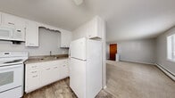 Located at 57 Whitehouse Dr in Rochester, NY, this single-floor unit offers a practical and well-organized layout encompassing a bedroom, bathroom, kitchen, living room, and hallway. The open kitchen and living area is a functional centerpiece of the home, featuring wood-look vinyl plank flooring, white cabinetry with decorative scalloped detailing, granite-look countertops, a double sink, and a full suite of white appliances including a refrigerator, electric range, and over-the-range microwave. The kitchen flows naturally into the carpeted living room, which is bathed in natural light from multiple windows offering outdoor views of the surrounding greenery. A wall-mounted air conditioning unit and baseboard heating provide year-round climate control. The spacious bedroom features soft carpeting, large windows that invite natural light, a built-in double-door closet, and a baseboard heater. Neutral gray walls throughout the unit create a clean, cohesive aesthetic. The bathroom is finished with classic tile surround in the tub/shower combination, offering a straightforward and functional design. A connecting hallway provides access to the bedroom and bathroom, completing this efficiently designed home.