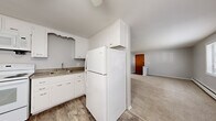 Located at 57 Whitehouse Dr in Rochester, NY, this single-floor unit offers a practical and well-organized layout encompassing a bedroom, bathroom, kitchen, living room, and hallway. The open kitchen and living area is a functional centerpiece of the home, featuring wood-look vinyl plank flooring, white cabinetry with decorative scalloped detailing, granite-look countertops, a double sink, and a full suite of white appliances including a refrigerator, electric range, and over-the-range microwave. The kitchen flows naturally into the carpeted living room, which is bathed in natural light from multiple windows offering outdoor views of the surrounding greenery. A wall-mounted air conditioning unit and baseboard heating provide year-round climate control. The spacious bedroom features soft carpeting, large windows that invite natural light, a built-in double-door closet, and a baseboard heater. Neutral gray walls throughout the unit create a clean, cohesive aesthetic. The bathroom is finished with classic tile surround in the tub/shower combination, offering a straightforward and functional design. A connecting hallway provides access to the bedroom and bathroom, completing this efficiently designed home.