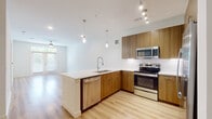 This well-appointed single-floor residence showcases a contemporary open-concept design that seamlessly connects the kitchen and living areas. The kitchen is a standout feature, boasting warm wood-toned cabinetry, crisp white quartz countertops, and a full suite of stainless steel appliances including a refrigerator with ice maker, electric range, built-in microwave, and dishwasher. A peninsula with an integrated sink and pendant lighting above adds both functionality and style, while a white mosaic tile backsplash and track lighting complete the modern aesthetic. Light hardwood-style flooring extends throughout the main living areas, creating a bright and cohesive feel. The spacious living area benefits from natural light streaming through large French doors that open to a green outdoor view, complemented by a ceiling fan overhead. The bedroom features plush carpeting, dual windows with ample natural light, and its own ceiling fan. The bathroom presents a clean, minimalist design with large-format tile flooring, a sleek vanity with white countertops and wood-toned cabinetry, and a glass-enclosed shower. Generous closet space and recessed lighting throughout further enhance the functionality of this thoughtfully designed residence.