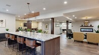 Located at 414 Andorra Glen Ct in Lafayette Hill, PA, this thoughtfully designed single-floor communal space offers a generous, open layout with a refined, contemporary aesthetic. Wide-plank wood-look flooring runs throughout, unifying the various areas under recessed lighting and decorative fixtures.

The kitchen and dining area anchors one end of the space, centered around a large, granite-topped island with dark wood cabinetry and leather bar stools. A wall-mounted television and natural light from multiple windows create an inviting atmosphere for gathering. Pendant lighting with a warm wood finish adds a stylish focal point above the island.

The living room features sophisticated lounge seating arranged around a glass-topped coffee table, anchored by a richly patterned area rug. A sleek linear electric fireplace is built into the wall, flanked by lush indoor plants and decorative shelving. A striking tiered chandelier with gold accents completes the look, while a large wall-mounted display screen enhances the entertainment experience.

A dedicated billiards area offers a full-size pool table beneath a modern multi-arm pendant light, with glass-partitioned walls providing a sense of openness. Structural columns with dark wood detailing add architectural character throughout the space, contributing to a polished and cohesive design that balances comfort with contemporary style.