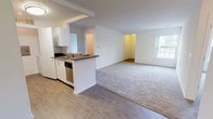 This single-floor, 2-bedroom, 2-bathroom unit features a thoughtfully designed open-concept layout that maximizes livability. The living and dining area is generously sized, with plush carpeting underfoot and natural light streaming in through windows. It connects seamlessly to the kitchen via a peninsula counter with a dark countertop, creating a natural divide while maintaining an open feel. The kitchen is equipped with stainless steel appliances, including a refrigerator, range, and dishwasher, complemented by white cabinetry and light wood-look flooring that extends throughout the kitchen and entry areas. Both bedrooms are well-proportioned, carpeted, and bright, each featuring windows that allow in natural light. One bedroom includes an en-suite bathroom, providing added privacy and convenience. The bathrooms are finished with tile flooring, white vanity cabinetry, and a tub-shower combination. The overall aesthetic is clean and neutral, with white walls throughout, creating a bright, move-in-ready environment. The layout, as seen in the dollhouse view, is compact yet efficiently organized, offering clear separation between living, sleeping, and kitchen zones.