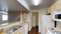 This single-floor residential space is designed with practicality and efficiency in mind. The kitchen serves as a central highlight, featuring a classic galley layout that maximizes functionality within a compact footprint. Light wood cabinetry lines the upper and lower walls, providing ample storage while lending a warm, neutral aesthetic to the space. The countertops are finished in a light tone that complements the cabinetry, and a double-basin sink is conveniently positioned along one side. White appliances, including a refrigerator, dishwasher, and range, offer a clean and cohesive look throughout the kitchen. Rich, dark hardwood flooring runs the length of the kitchen, adding depth and a touch of elegance that contrasts nicely with the lighter tones of the cabinetry and walls. Overhead, a flush fluorescent light fixture provides bright, even illumination across the workspace. A doorway at the end of the kitchen connects to an additional room or hallway, suggesting thoughtful flow between spaces. The walls are finished in a soft, neutral white that keeps the interior feeling open and inviting. Overall, the space presents a clean, well-maintained environment with a functional design well-suited for everyday living.