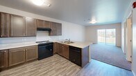 Located at 1410 Catron Blvd in Rapid City, South Dakota, this single-floor, one-bedroom, two-bathroom residence offers a thoughtfully designed layout that maximizes every square foot. The open-concept kitchen and living area serves as the heart of the home, featuring rich dark cabinetry, light countertops, a peninsula with a double sink, and black appliances including a range and dishwasher. Wood-look luxury vinyl flooring runs throughout the main living areas, creating a clean and cohesive aesthetic. The kitchen flows naturally into the carpeted living area, which benefits from a sliding glass door that invites natural light and outdoor views. The bedroom is a comfortable, carpeted retreat with a window overlooking the surrounding greenery, offering a quiet and private atmosphere. Both bathrooms are designed with accessibility in mind, featuring wall-mounted vanities, large mirrors, grab bars, and open roll-in shower areas with wood-look flooring. A dedicated laundry room houses a full-size front-loading washer and dryer, adding everyday convenience. The overall design is clean, modern, and functional, with a neutral color palette and practical finishes throughout.