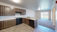 Located at 1410 Catron Blvd in Rapid City, South Dakota, this single-floor, one-bedroom, two-bathroom residence offers a thoughtfully designed layout that maximizes every square foot. The open-concept kitchen and living area serves as the heart of the home, featuring rich dark cabinetry, light countertops, a peninsula with a double sink, and black appliances including a range and dishwasher. Wood-look luxury vinyl flooring runs throughout the main living areas, creating a clean and cohesive aesthetic. The kitchen flows naturally into the carpeted living area, which benefits from a sliding glass door that invites natural light and outdoor views. The bedroom is a comfortable, carpeted retreat with a window overlooking the surrounding greenery, offering a quiet and private atmosphere. Both bathrooms are designed with accessibility in mind, featuring wall-mounted vanities, large mirrors, grab bars, and open roll-in shower areas with wood-look flooring. A dedicated laundry room houses a full-size front-loading washer and dryer, adding everyday convenience. The overall design is clean, modern, and functional, with a neutral color palette and practical finishes throughout.