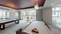 Located on South Boulevard in Charlotte, NC, this single-floor amenity space at Uptown 550 is thoughtfully designed to offer residents a variety of recreational and social environments. The entrance foyer greets visitors with large floor-to-ceiling windows that flood the space with natural light, polished large-format tile flooring, a sleek curved black velvet sofa, a glass coffee table, and an elegant crystal chandelier overhead.

The billiards lounge is a standout feature, accommodating multiple full-sized pool tables beneath a dramatic deep burgundy tray ceiling. Stylish pendant lights hang above, and floor-to-ceiling glass doors open to an outdoor terrace furnished with high-top tables and chairs, providing views of the surrounding urban streetscape.

Adjacent to the billiards area, a dedicated golf simulator room houses a full Full Swing simulator setup, complete with a synthetic turf hitting mat and projection screen, ideal for year-round play. A comfortable lounge seating area nearby, finished with hardwood-style flooring and large windows overlooking the city, offers plush barrel chairs and a glass coffee table — perfect for relaxing before or after a round.

A media lounge features dual wall-mounted flat-screen televisions and casual seating for watching sports and entertainment. The space also includes a well-appointed wet bar with white cabinetry, a sink, wine refrigerator, and decorative geometric pendant lighting, making it ideal for hosting social gatherings.