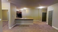 This single-floor space features an open-concept design that seamlessly connects the kitchen and living area, creating a cohesive and functional layout. The kitchen is equipped with dark-toned cabinetry, black appliances including a refrigerator, and a peninsula that doubles as a workspace and casual dining surface. The peninsula features a granite-style countertop and an integrated sink, making it a practical focal point that separates the kitchen from the adjoining living area.

The living area is finished with wall-to-wall carpeting in a neutral tone, providing a warm and comfortable feel underfoot. Walls throughout are painted in a soft, muted yellow-green hue that lends a calm, understated ambiance to the space. Flush-mount ceiling lights illuminate both zones evenly, ensuring adequate brightness throughout.

A dark-paneled entry door is visible along the far wall, providing a defined point of entry. White baseboards trim the perimeter of the space, offering a clean contrast to the carpeted floors. Overall, the layout prioritizes efficiency and flow, making the space well-suited for everyday living.