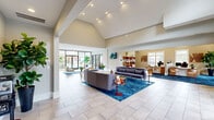 Located at 7305 Village Center Blvd in Fairburn, Georgia, this single-floor community clubhouse offers a generous and thoughtfully designed gathering space. The architecture is defined by dramatic vaulted ceilings with exposed beam detailing, recessed lighting, and large floor-to-ceiling windows and glass doors that flood the interior with natural light and provide views of the outdoor pool area.

The expansive living room serves as the heart of the space, featuring large-format tile flooring, bold blue area rugs, and modern furnishings arranged to encourage both relaxation and socializing. A statement sliding barn door adds a rustic-modern contrast to the otherwise contemporary interior, while lush potted plants contribute a vibrant, welcoming atmosphere.

A dedicated office area is set apart with rich blue carpet, warm wood open shelving, a workstation desk, and ample natural light from surrounding windows — ideal for administrative or leasing functions. The kitchen is compact yet functional, showcasing white cabinetry, granite-style countertops, pendant lighting, and a striking horizontal wood plank accent wall that adds warmth and texture. A built-in beverage cooler and sink complete the setup.

Multiple entry points, including glass-paneled front doors, seamlessly connect the interior to the outdoor surroundings, enhancing the overall sense of openness throughout the space.