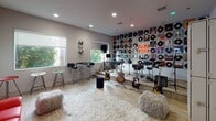 The Lyric LP Lounge is a single-floor music-themed space designed for both relaxation and creative expression. The main area is divided into two distinct zones that flow seamlessly into one another. The lounge zone features white-painted brick walls, light wood flooring, and a bold red leather sofa as its centerpiece, accompanied by a cylindrical ottoman and additional bar-height seating. Two wall-mounted flat-screen TVs are positioned for easy viewing, while a large window allows natural light to fill the room, creating a comfortable and inviting atmosphere. A glass-paneled entry door adds a modern, polished touch to the entrance. The music zone commands immediate attention with its dramatic feature wall — an expansive grid display of vinyl LP records covering the surface from floor to ceiling, punctuated by a glowing neon sign that reads "Lyric LP Lounge." The instrument collection is impressive, including multiple electric and acoustic guitars on stands, a full electronic drum kit, a keyboard, microphone stands, and amplifiers, making the space fully equipped for live performance or jam sessions. A plush white area rug and fluffy floor cushions add warmth and texture, while recessed lighting and additional speakers complete the well-appointed setup. This space is ideal for music lovers, performers, and anyone seeking an immersive, creative environment.